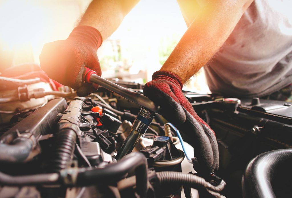 The red gloved hand of the auto mechanic is fastening the bolt with the socket wrench to fix the vehicle engine, sunlight on background